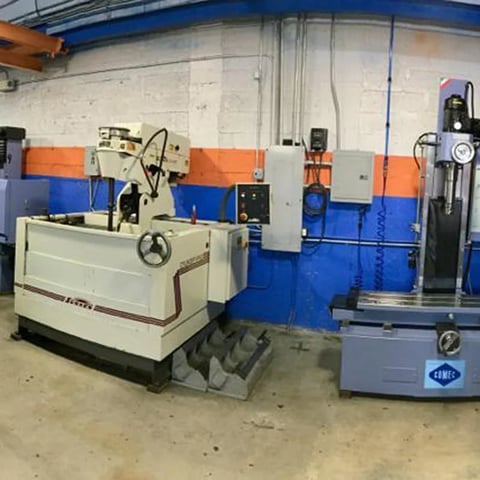 Industrial workshop with white and cream CNC machine on left, blue machine on right, orange and blue wall panels, and overhead crane on concrete floor